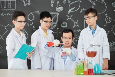 Laboratuvar önlüğü ve gözlük takmış meraklı Vietnamlı bir grup öğrenci deney tüpünde reaktörleri karıştıran öğretmenlerine bakıyor.