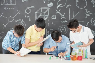 Okul öğrencileri yanlarında durup ders kitaplarına yazarken petri kabındaki bakterilere mikroskopla bakıyor.