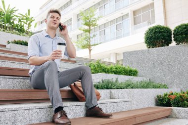Genç ve mutlu girişimci basamaklarda oturuyor, kahve içiyor ve telefonla arıyor.