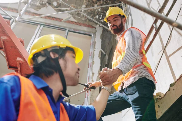 建設中の建物の高い階段を登るために同僚を助ける深刻なひげそりのビルダー — ストック写真