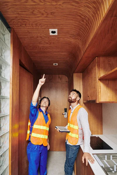キッチンエリアの木製の天井に設置したライトソケットを手にデジタルタブレットを持つフォアマンに示す電気技師 — ストック写真