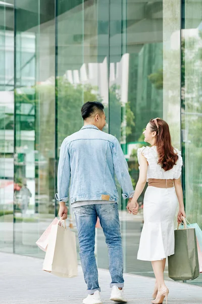 Happy young Asian couple with paper-bags walking along the street with stores and boutiques, view from the back