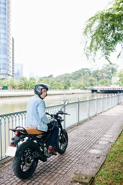 一个头戴安全帽的严肃的亚洲人骑着摩托车沿着河岸向前看 — 图库照片
