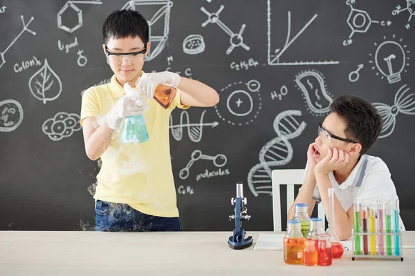 Excited schoolboy in goggles opening mouth when looking at his classmate mixing smoking colorful chemical liquids
