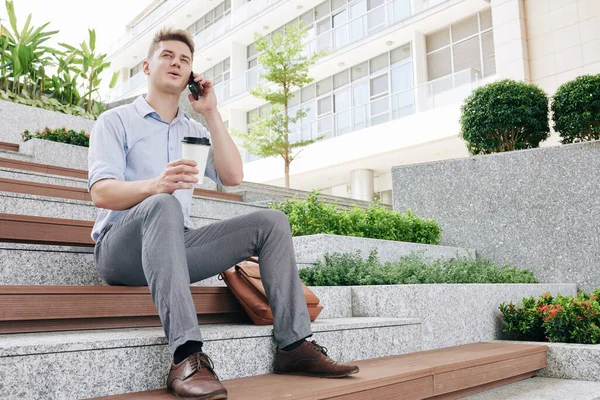 Genç ve mutlu girişimci basamaklarda oturuyor, kahve içiyor ve telefonla arıyor.