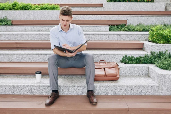 Ciddi genç işadamı klasördeki belgeleri okuyor, mali raporları kontrol ediyor ve düzeltmeler yapıyor.