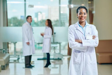 Klinikte duran beyaz önlüklü, gülümseyen genç siyahi doktorun portresi.