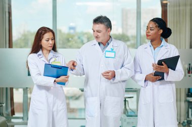 Hastane koridorunda yürürken stajyer doktorların teşhis koymasına yardım eden olgun bir doktor.