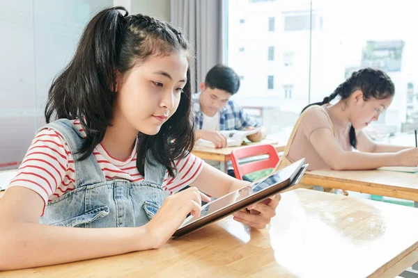 认真的亚洲女学生坐在课桌前阅读平板电脑上的文字 — 图库照片