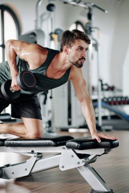 Ciddi sakallı, formda bir adam. Yedek kulübesine yaslanıyor ve modern spor salonunda tek kollu halterli sıra yapıyor.