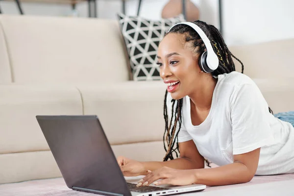 Excited beautiful young Black woman in dreadlocks lying on the floor and studying or working on laptop