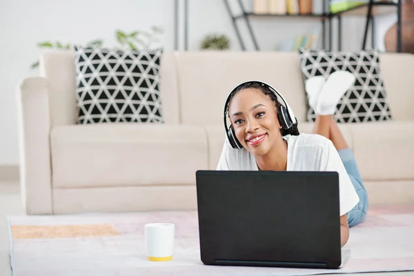 正面黑人妇女躺在地板上和在家里用笔记本电脑工作时戴耳机的画像 — 图库照片