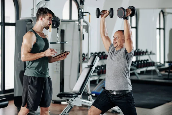 Trener Fitness Robi Notatki Dokumencie Tablecie Gdy Jego Klient Podnosi — Zdjęcie stockowe