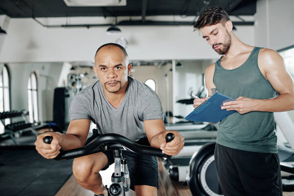 Mature mixed-race man riding fast on stationary bicycle in gym, his personal trainer taking notes in document