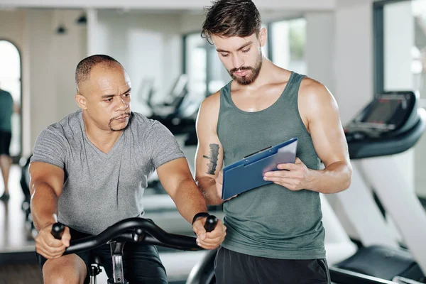 Fit Man Diskuterar Träningsplan Med Sin Tränare När Rider Stationär — Stockfoto