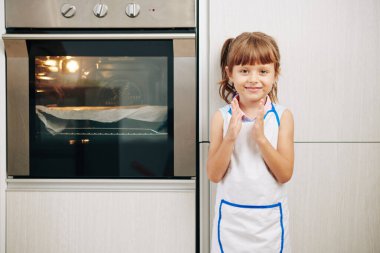 Mutfakta fırında durup pasta pişirilmesini bekleyen gülümseyen kızı alkışlıyor.