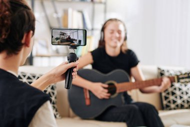 Genç çocuk, kız kardeşini ya da arkadaşını gitar çalarken ve şarkı söylerken çekerken Monopod 'da akıllı telefon kullanıyor.