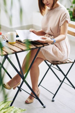 Kafe masasında bir fincan kahveyle oturan ve uzaktan öğrenme üzerine kitap okuyan Asyalı genç bir kadının kırpılmış görüntüsü.