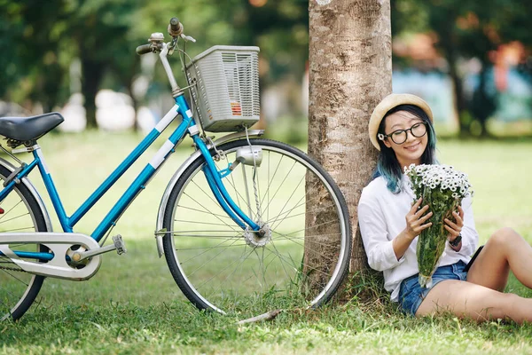 幸せな美しい若い中国人女性座って下の木の横に彼女の自転車と彼女の手の中に白い花の花束を見て — ストック写真