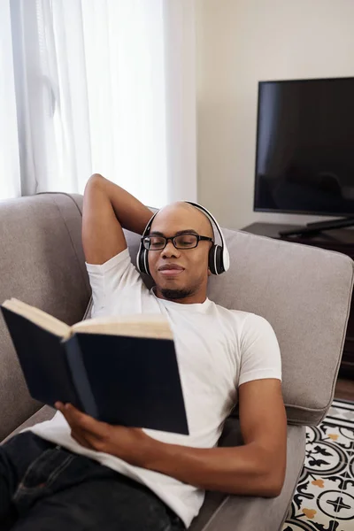 Sonriente Joven Hombre Negro Los Auriculares Relajante Sofá Lectura Novela —  Fotos de Stock