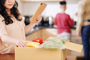 Tablet bilgisayarlı ev hanımı kocası ve küçük oğlu arka planda yemek pişirirken büyük karton kutudan taze yiyecek alıyor.