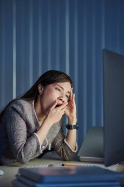 Genç Vietnamlı iş kadını bilgisayar ekranındaki e-posta belgesini okurken esniyor, karanlık ofiste çalışıyor.
