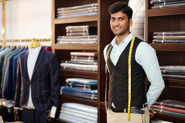 Portrait of handsome smiling Indian tailor with measuring tape around his neck standing in atelier and looking at camera