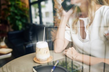 Restoranda nefis buzlu kahve içen ve telefonla arayan genç bir kadının görüntüsü.