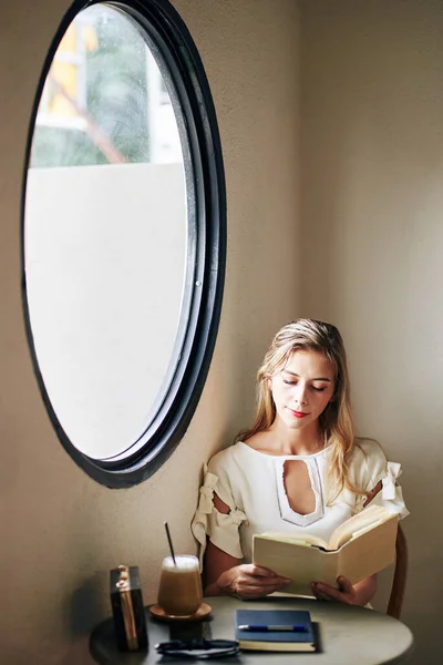 Sjarmerende Smilende Ung Kvinne Som Sitter Ved Bordet Leser Interessant – stockfoto