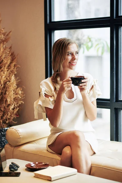 一个非常快乐的年轻女子喝着一杯美味的咖啡从窗户往外看的画像 — 图库照片