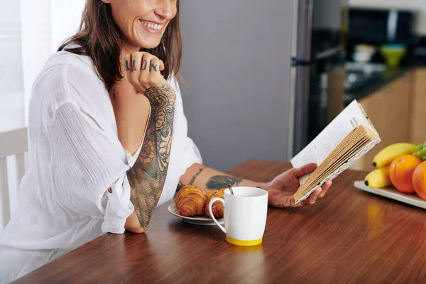 幸せな若い女性読書面白い本を食べるときクロワッサンとコーヒーを飲むカップのための朝食 — ストック写真