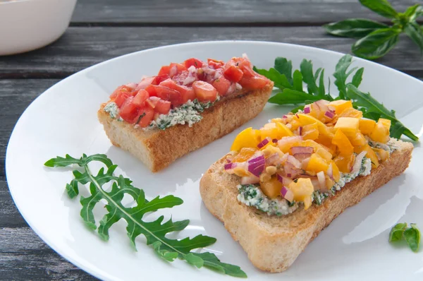 Bruschetta with cheese red and yellow tomatoes on a white plate. The composition is decorated with leaves of basil, arugula, pepper and vegetables. Side view on an old wooden background. Blurred.