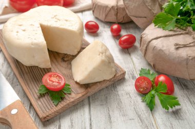 Adighe peyniri üçgen bir parçayı kesmiş. Tahtada peynir var. Domateslerin yanında maydanoz ve dereotu var. Hafif ahşap zemin. Yakın plan..