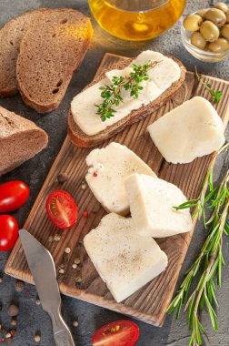 Adighe peyniri parçalara ayrılmış. Tahtada peynir var. Kekik, biberiye, baharat ve vişneli domatesle servis edilir. Arka planda zeytin, çavdar ekmeği ve zeytinyağı var. Arka plan gri beton. Yukarıdan görüntüle.