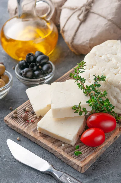 Adyghe cheese is cut into pieces. Cheese on a wooden board. Garnished with thyme, spices and cherry tomatoes. In the background olives, packaged cheese and olive oil. Background gray concrete. Close-up. — Stock Image Adyghe Cheese Cut Pieces Cheese Wooden Board Garnished Thyme Spices Stock Image