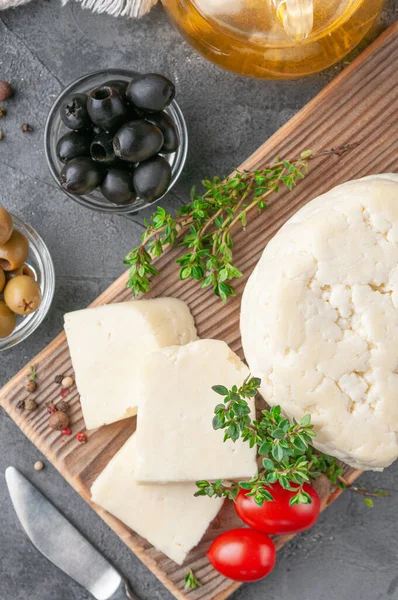 Cut Adyghe cheese into pieces. Cheese on a wooden board. Served with thyme, spices and cherry tomatoes. In the background are olives, packaged cheese and olive oil. Background gray concrete. View from above. — Stock Image Cut Adyghe Cheese Pieces Cheese Wooden Board Served Thyme Spices Stock Photo
