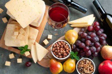 Paketlemeden yuvarlak peynirli. Peynirin yanında beyaz bir tabakta şarap, üzüm ve armut şişeleri var. Ön planda bir bardak kırmızı şarap, dilimlenmiş armut ve peynir var. Logo için bir yer. Gri keten arka plan