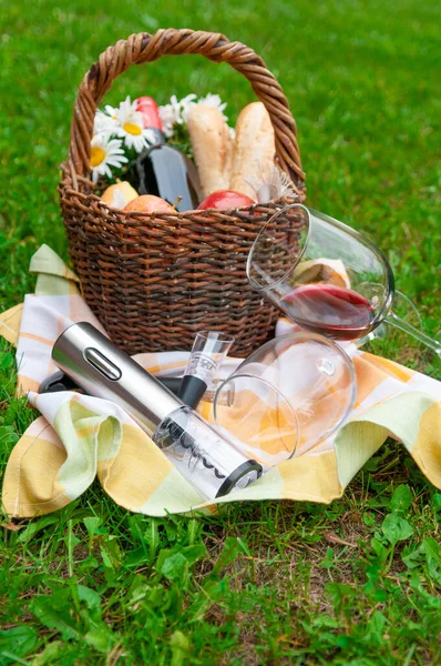 An electric corkscrew lies on a towel. Near the aerator for wine and two glasses of wine. In the background is a basket of picnic products. A basket is standing on the green grass. Close-up