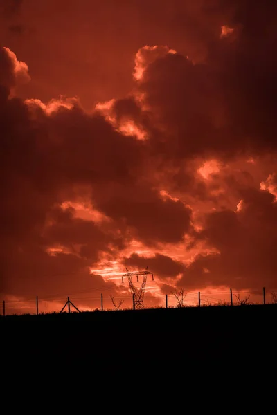 Scary sunset, with red sky and clouds, after the storm. Power pylons, cell towers and metal structures behind wired fences. Pollution, in a devastated landscape. Evil environment.