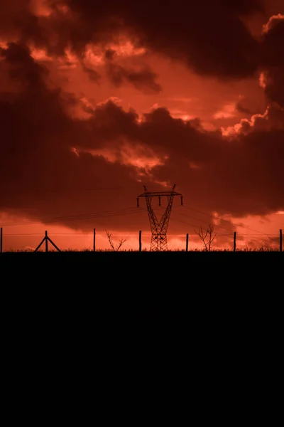 Scary sunset, with red sky and clouds, after the storm. Power pylons, cell towers and metal structures behind wired fences. Pollution, in a devastated landscape. Evil environment.