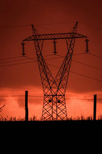 Scary sunset, with red sky and clouds, after the storm. Power pylons, cell towers and metal structures behind wired fences. Pollution, in a devastated landscape. Evil environment.
