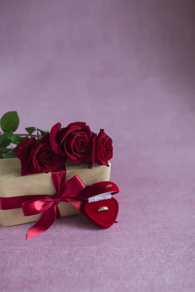 red roses on a gift box, a box with a ring lies on a pink background, flowers, a gift and a ring on a pink background, a proposal to get married (vertically, copy space)
