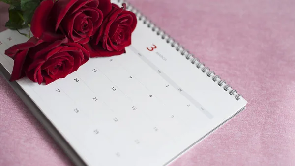 the calendar for March and three red roses lie on a pink background, a calendar with numbers lies on a pink background, red roses lie on the calendar
