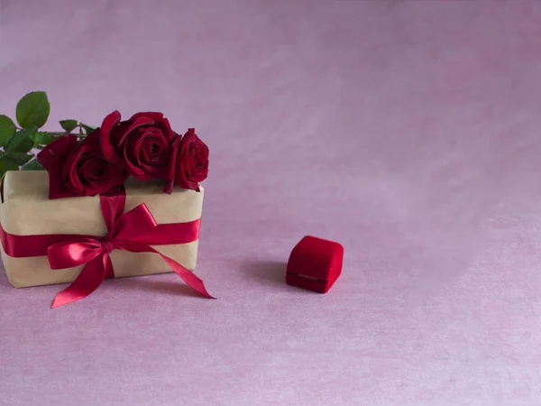 three red roses, and red box with engagement ring (closed) box tied with red ribbon on pink background