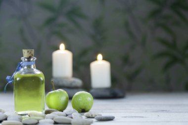 şişe elma yağı masaj, nehir çakıl, iki küçük yeşil elma ve ahşap masa ve bitkisel arka plan üzerinde iki yanan mumlar