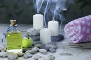 şişe elma yağı masajı, nehir çakıl taşları, küçük bir yeşil elma, havlu ve duman ahşap masa ve bitkisel arka plan ile üç mum 