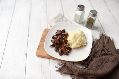 Beyaz ahşap zemin üzerinde susamlı biftek tabağı patates püresi ve baharatlarla servis edilir.