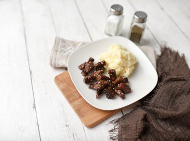 Beyaz ahşap zemin üzerinde susamlı biftek tabağı patates püresi ve baharatlarla servis edilir.