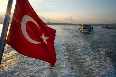 Kırmızı Türk bayrağının vapurda su ve dalgalara karşı yakın olduğu deniz Boğazı (boğaz) gün batımı üzerinde. Diğer yolcu gemisine odaklanın. Bayrağın üzerinde güneş patlaması var. Istanbul, Türkiye