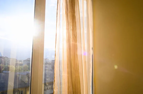 Güneş perdeler ve pencereden parlıyor. Gün batımında güneşli bir daire. Şehir manzarası ve mavi gökyüzü.